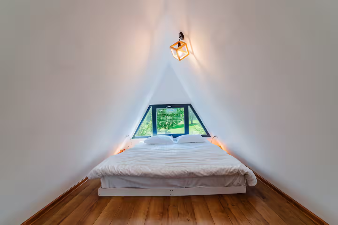 Dormitor la mansarda cabanei Stoic A-frame, cu fereastră triunghiulară spre natură Dormitor la mansarda cabanei Stoic A-frame, cu fereastră triunghiulară spre natură