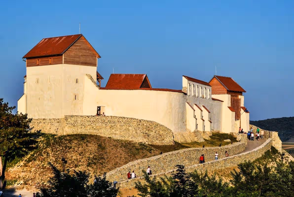 Cetatea Feldioara pe deal, zidurile albe și acoperișurile roșii la apus Cetatea Feldioara pe deal, zidurile albe și acoperișurile roșii la apus