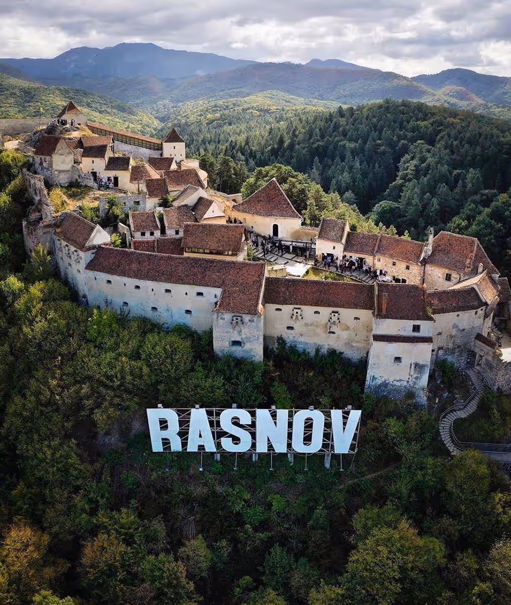 Cetatea Râșnov văzută din dronă, cu literele RÂȘNOV și Munții Bucegi în fundal Cetatea Râșnov văzută din dronă, cu literele RÂȘNOV și Munții Bucegi în fundal