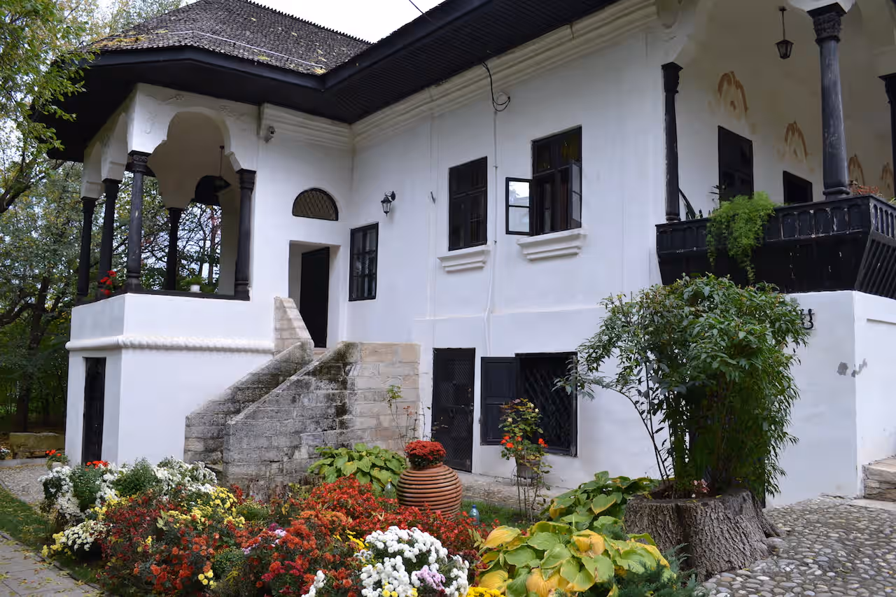 Fațada albă a Conacului Bellu cu scară exterioară și grădină cu flori Fațada albă a Conacului Bellu cu scară exterioară și grădină cu flori